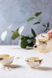 从茶壶里倒入热绿茶，放在两个传统的中国粘土陶瓷杯上，站在白色大理石桌上。 背景上有绿色的树枝。