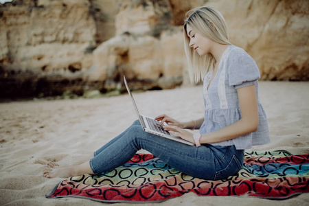 金发的年轻女子，穿着休闲装，手提电脑坐在海边