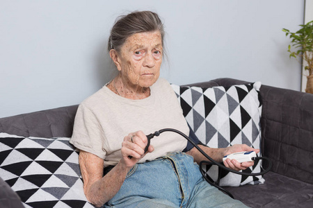 话题是很老的人和健康问题。一位白人女性，90岁，有皱纹和白发，坐在沙发上，用血压监测仪测量血压。