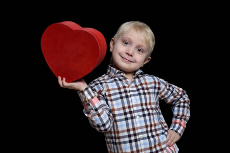 微笑的金发男孩穿着格子衬衫，手里拿着一个红色的心形盒子。 黑色背景的隔离物
