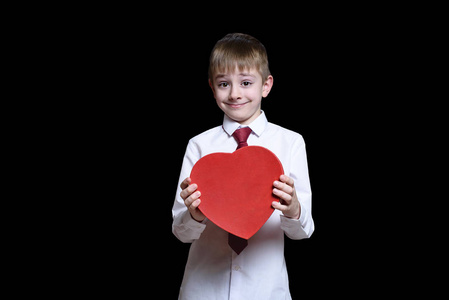 漂亮的头发男孩穿着衬衫和领带，手里拿着一个红色的心形盒子。 爱和家庭观念。 黑色背景的隔离物
