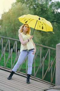 晚上，穿着黄色衬衫和牛仔裤的可爱女孩带着一把明亮的雨伞在桥上散步