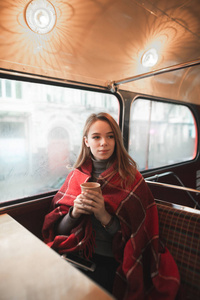 冬天的肖像，一个年轻的女孩覆盖着一条毯子，坐在窗户附近的一杯咖啡，看着一边。 一个可爱的年轻女孩在咖啡店喝咖啡的舒适照片。