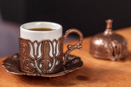 装饰丰富的小传统土耳其杯，用于木制背景上的浓黑咖啡。 咖啡馆的咖啡概念。 土耳其咖啡在金属浮雕铜杯上的木制桌子上。