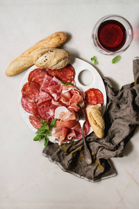 在白色陶瓷板上用面包和一杯红酒铺在白色大理石背景上的布上，用Jamon Salami Chorizo香肠切片的反胃肉盘。 平躺空
