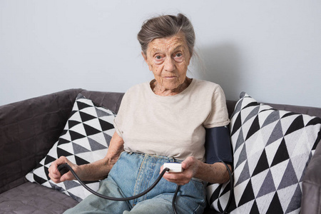 这个话题是很老的人和健康问题。 一位90岁的高加索老年妇女，长着皱纹和白发，坐在沙发上，用血压监测仪测量血压。