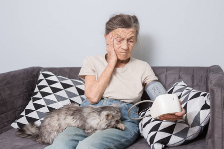 这个话题是很老的人和健康问题。 一位90岁的高白种人妇女，长着皱纹和白发，坐在沙发上，带着宠物猫，用眼压计。 高血压头痛。