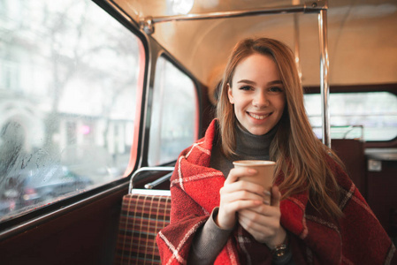 一个微笑的女孩的舒适肖像，上面覆盖着一个格子，手里拿着一杯咖啡，坐在咖啡馆里看着相机。 在咖啡店里，坐在桌子旁喝咖啡的可爱女孩