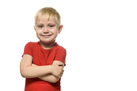 微笑的金发小男孩穿着一件红色T恤，双臂交叉站立。 在白色背景下分离