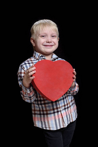 微笑的金发男孩穿着格子衬衫，手里拿着一个红色的心形盒子。 在黑色背景上隔离。 垂直框架