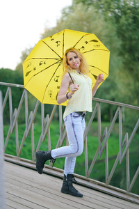 晚上，穿着黄色衬衫和牛仔裤的可爱女孩带着一把明亮的雨伞在桥上散步
