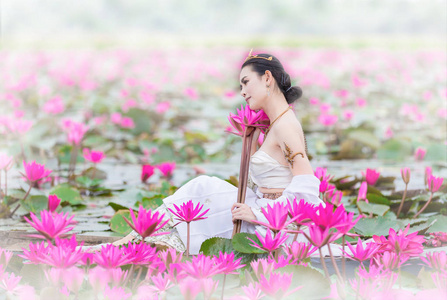 美丽而幸福的泰国女人穿着白色的当地传统服装，名字叫纳基，坐在红莲花田池塘里的木船上