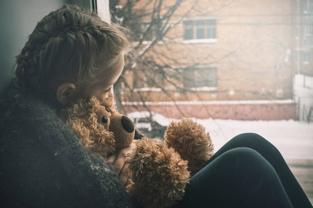 婴儿情感。 带着泰迪熊的小女孩坐在窗台上看着窗外。 孩子正在等父母。