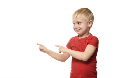 微笑的金发小男孩穿着一件红色衬衫站着，食指对侧。 在白色背景下分离