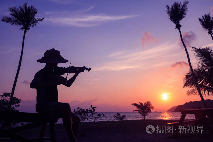 当地音乐家亚洲男子拉小提琴在椰子海滩日出剪影艺术家在紫色的天空背景。