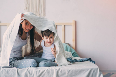 快乐的母子在舒适的周末早晨在床上玩，躲在毯子下。 休闲生活方式，真正的家庭