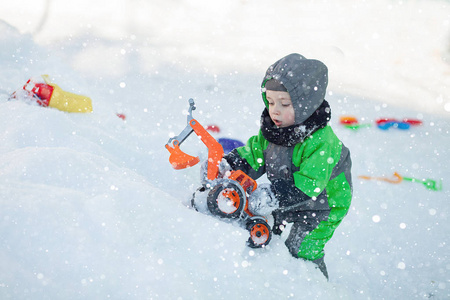 可爱的小蹒跚学步的孩子坐在雪地上，在公园里玩他的黄色拖拉机玩具的肖像。 孩子们在户外玩耍。 带建筑玩具的快乐男孩。 生活方式的概