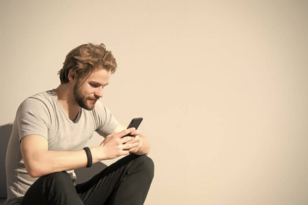 大胡子男子用手机在灰色墙上。有男子气概的智能手机在阳光明媚的户外。有胡子和金发的家伙在休闲 t恤短信短信。新技术与现代生活, 复