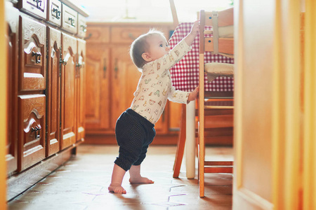 女婴站在厨房的地板上，拿着家具。 小孩子在家里大吵大闹