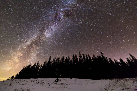 冬山夜景全景。 银河明亮的星座在深蓝色的星空上，在黑暗的云杉松树上，森林在日落后的地平线上柔和地发光。 广角射击。