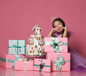 小亚洲女孩在皇冠和紫色礼服与许多礼物礼物箱子和生姜饼干蛋糕生日庆祝