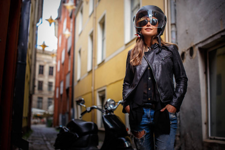 穿着防护头盔太阳镜皮夹夹克穿着撕破牛仔裤的时尚女孩, 站在一条狭窄的老街道上的黑色摩托车旁