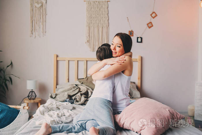 快乐的母亲早上拥抱她蹒跚学步的儿子坐在家里的床上休闲生活家庭生活