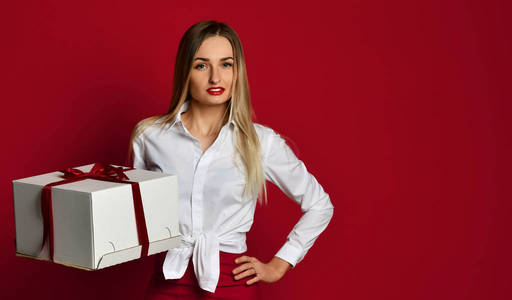 年轻的金发碧眼的妇女拿着白色的盒子礼物礼物微笑地尝试打开它在生日党庆祝在红色