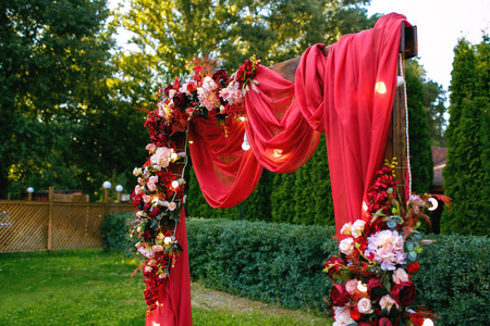 公园里的绿草上矗立着鲜花和绿色植物的红色婚礼拱门.照片