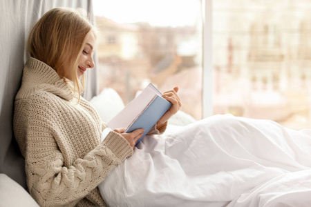 美丽的年轻女子穿着针织毛衣，坐在家门口看书。 冬季气氛
