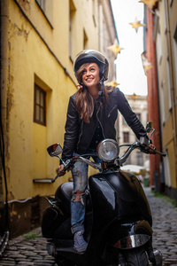 在欧洲古老狭窄的街道上, 穿着皮夹克穿着撕破牛仔裤的快乐嬉皮士女孩穿着黑色经典摩托车