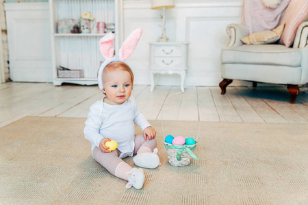 复活节时戴兔子耳朵的小女孩。女孩拿着篮子和彩蛋坐在家里的地板上，玩复活节彩蛋狩猎。复活节节日快乐庆祝春天