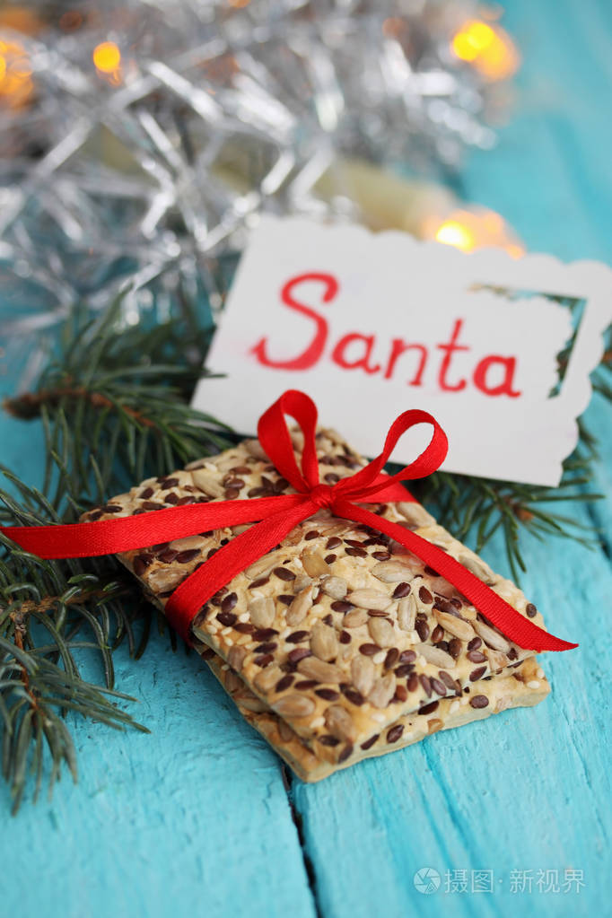 带有亚麻籽和向日葵的饼干，上面写着圣诞老人的卡片。 木制背景上的礼物。 带有复制空间的圣诞背景。