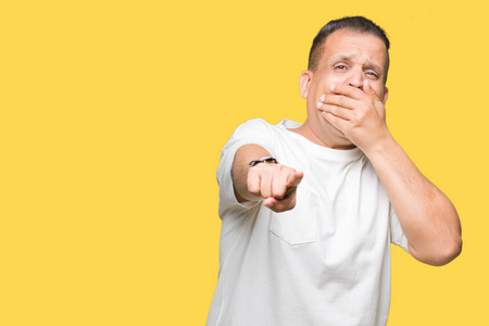 中年阿拉伯男人穿白色t恤在孤立的背景上笑你指着相机用手指，用手捂住嘴羞愧的表情