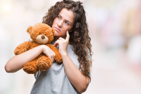 年轻的黑发女孩抱着玩具熊在孤立的背景下严肃的脸思考问题非常困惑的想法