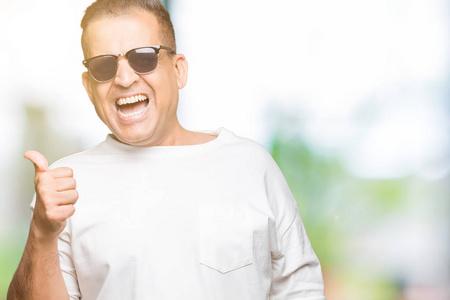 中年阿拉伯男人穿着白色T恤和太阳镜，在孤立的背景上微笑，快乐的脸看着，用拇指指向一边。