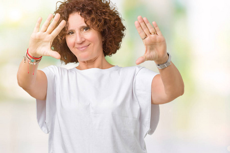 漂亮的中老年妇女，穿着白色T恤，在孤立的背景下微笑，用手手掌和手指相机透视框架