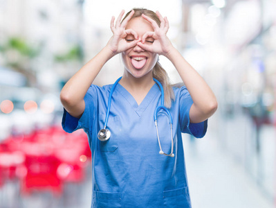 年轻的金发外科医生，女医生，在孤立的背景下做着好的手势，比如双筒望远镜伸出舌头，眼睛透过手指看。疯狂的表情。