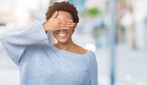 年轻美丽的非洲裔美国妇女穿着一件毛衣，在孤立的背景上微笑和笑，用手捂住眼睛，以获得惊喜。盲目的观念。