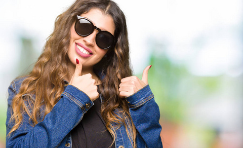 年轻漂亮的女人戴着太阳镜，用手做快乐的竖起大拇指的手势。赞许的表情看着相机，显示出成功。