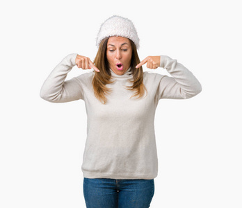 美丽的中年妇女穿着冬天的毛衣和帽子，戴在孤立的背景上，手指向下指向广告，惊讶的脸和张开的嘴
