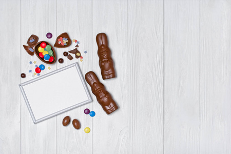 空白白框传统复活节糖果模型的顶部视图，如巧克力蛋和白色木制背景上明亮的彩色悬垂