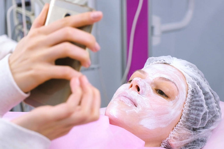 女人在手机里读着一些东西躺在美容师办公室里的程序上, 脸上戴着面具