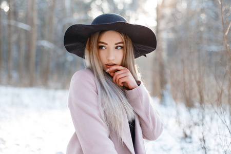 令人惊叹的迷人，漂亮的年轻，戴着豪华的黑色帽子，穿着时尚的粉红色温暖外套，在冬日公园的阳光明媚的日子里摆姿势。时髦的女孩散步