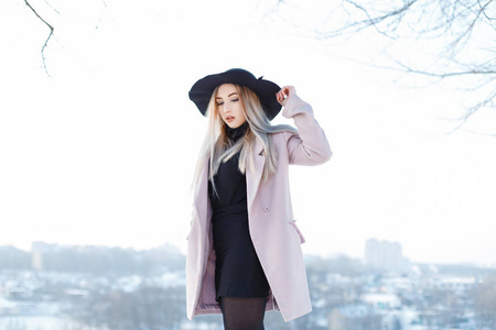 时尚迷人，优雅的年轻女人，穿着粉红色的外套，戴着黑色的帽子，穿着针织连衣裙，在白色的冬日天空中摆姿势。迷人的女孩在冬天散步。