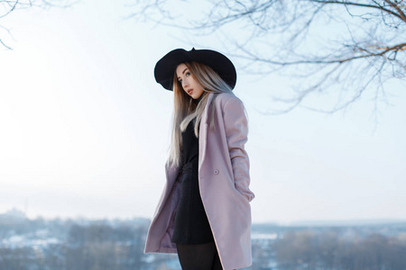 时尚美丽的年轻女人，金发，戴着一顶老式别致的黑色帽子，穿着一件粉红色的优雅外套，在户外对着冬天的天空摆姿势。迷人的时尚女孩在大自