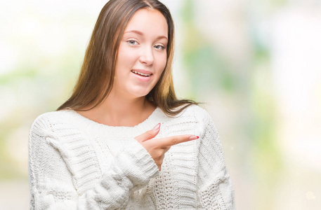 年轻漂亮的白种人女人穿着冬天的毛衣，在孤立的背景上欢快地笑着，脸上的微笑用手和手指指向一边，脸上的表情快乐而自然地看着镜头。