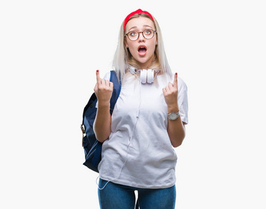 年轻的金发学生女人戴着眼镜和背包在孤立的背景上惊讶和惊讶地抬头，用手指和举起的手臂指着。