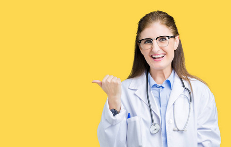 中年成熟的医生女人穿着医用外套，在孤立的背景上微笑，快乐的脸看着，用拇指指向一边。