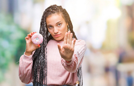 年轻的非裔美国女孩吃粉红色甜甜圈在孤立的背景上，张开手做停止标志，严肃和自信的表情防御手势